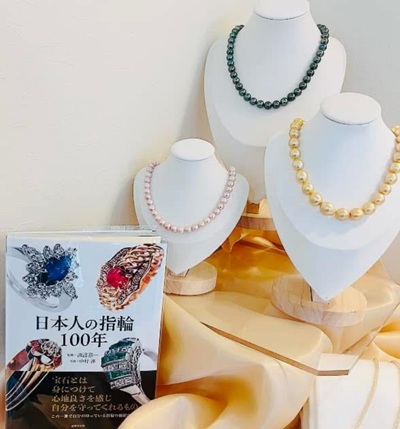 書籍『日本人の指輪100年』と、白色トルソーにディスプレイされた3本のパールネックレス(上品な淡いピンク系パール、シックなタヒチ黒蝶パール、輝くゴールド系南洋真珠)。柔らかなクリーム色のクロスの上に美しく飾られています。